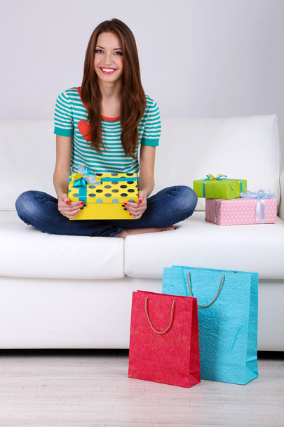 Beautiful young woman sitting on sofa with gift box on gray background - 写真・画像