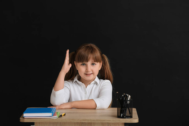 Pieni oppilas, jolla on kohotettu käsi, istuu koulun pöydällä tummaa taustaa vasten - Valokuva, kuva