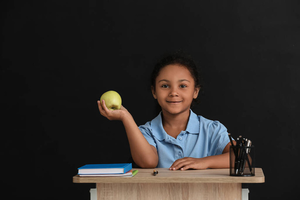 Little African-American pupil sitting at school desk against dark background - Foto, afbeelding