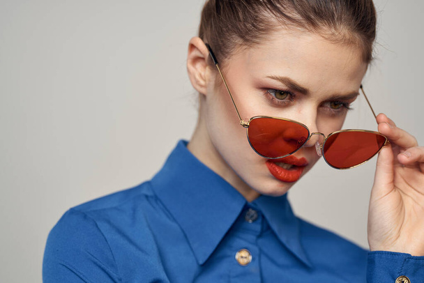 Fashionable woman in sunglasses and a blue shirt gesturing with her hands on a light background cropped view - Photo, Image