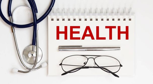 stethoscope,glasses and pen with notepad with text HEALTH on white background - Φωτογραφία, εικόνα