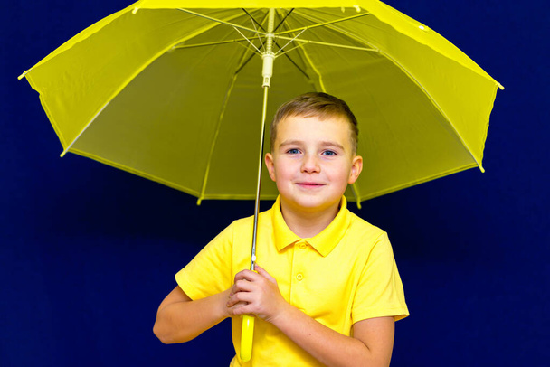 Attraktiv überrascht kaukasischen blonden Schuljungen mit Regenschirm auf blauem Studiohintergrund.Junge zeigt alle Arten von Fratzen. - Foto, Bild