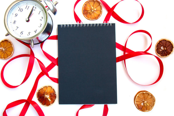 Φωτεινή εορταστική κόκκινη κορδέλα και μπούκλα και ένα σημειωματάριο με μαύρες σελίδες, φέτες αποξηραμένου λεμονιού και vintage ασημένιο ξυπνητήρι που απομονώνονται σε λευκό φόντο. Λίστα στόχων για το νέο έτος. Κενό πρότυπο - Φωτογραφία, εικόνα