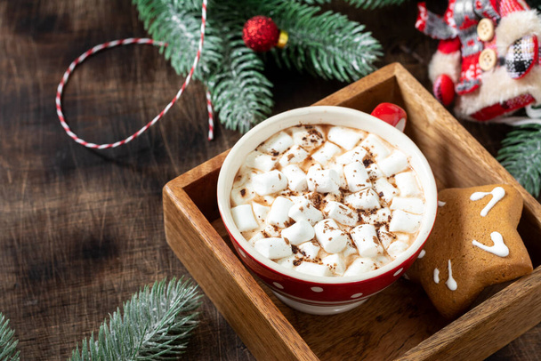 Cocoa with marshmallows in a red Cup and gingerbread on a tray on a Christmas background. Red decorations on spruce branches on a brown background - Foto, afbeelding