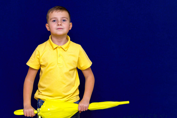 Attractive surprised caucasian blond schoolboy holds unexpanded umbrella on blue studio background.Boy shows all kinds of grimaces. - Photo, image