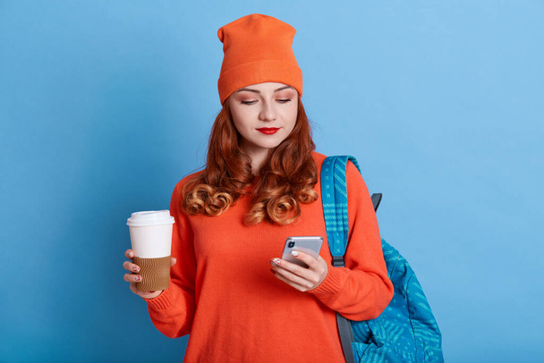 Photo of young woman feels good from received email, holds coffee to go, has backpack, carries rucksack, stands against blue wall, looks at device screen, wearing casual attire. - Fotó, kép