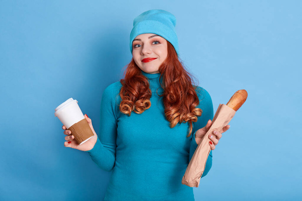 Puzzled woman shrugs shoulders, spreads hands with take away coffee and long loaf, wears casual clothes, posing against blue background, ginger girl has snack. - Fotoğraf, Görsel