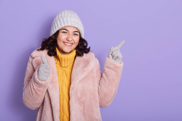 Smiling young brunette woman yellow sweater, pink fur coat , gray cap and mittens, posing isolated on lilac wall, pointing index finger aside and shows thumb up with other hand. - Zdjęcie, obraz