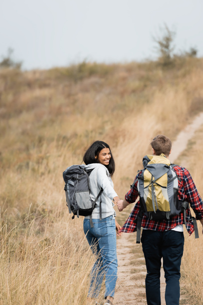Smiling african american woman with backpack holding hand of boyfriend while hiking on path  - Фото, изображение