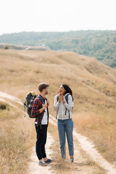 Cheerful african american woman standing near boyfriend with backpack on path during trip  - Zdjęcie, obraz