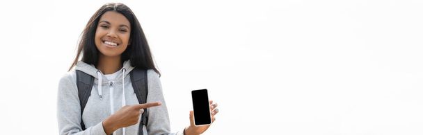 Usmívající se africký americký turista ukazuje prstem na smartphone s prázdnou obrazovkou izolované na bílé, banner  - Fotografie, Obrázek