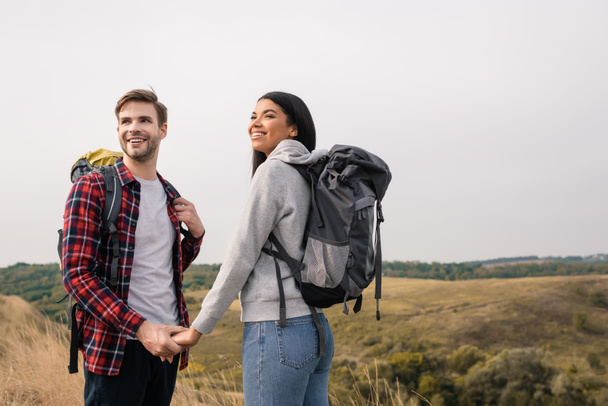 Sonriente pareja multiétnica con mochilas cogidas de la mano con colinas sobre un fondo borroso  - Foto, imagen