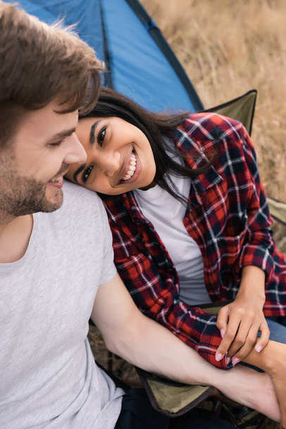 Smiling african american woman looking at boyfriend on blurred foreground during camping on lawn  - Фото, изображение