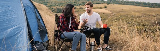 Sonriente pareja multiétnica con taza y termos hablando cerca de la tienda durante el camping, pancarta  - Foto, imagen
