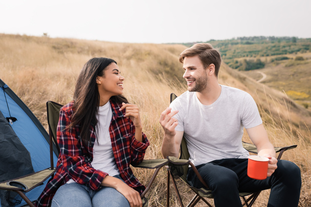 Smiling interracial couple with cup talking on chairs near tent during trip  - Фото, зображення