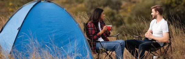 Smiling interracial couple with cup sitting on chairs near tent during camping, banner  - Foto, Imagen
