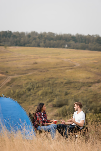 Man talking to smiling african american girlfriend with cup during camping near tent with field on blurred background  - Foto, Imagem