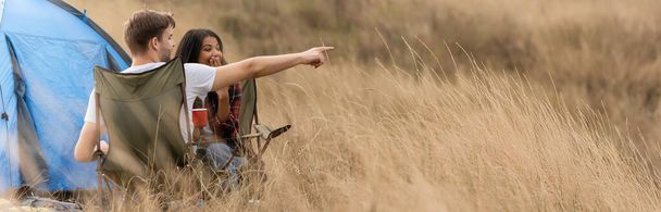Mężczyzna wskazując palcem w pobliżu afrykańskiej dziewczyny z filiżanką podczas kempingu na trawniku, sztandar  - Zdjęcie, obraz