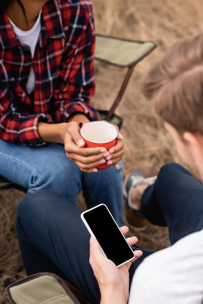 Hombre sosteniendo teléfono inteligente con pantalla en blanco cerca de mujer afroamericana con taza durante el camping  - Foto, imagen