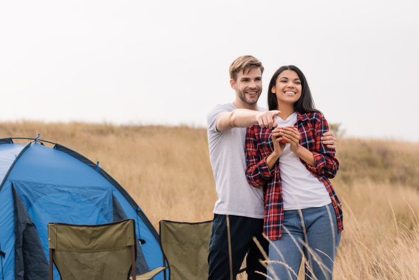 Smiling man embracing african american girlfriend with cup and pointing with finger during camping on lawn  - Photo, Image