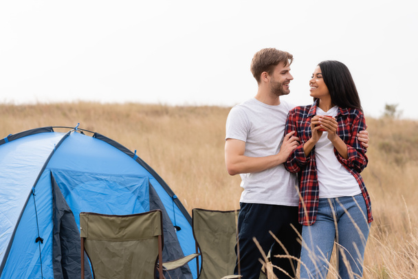 Smiling man hugging african american girlfriend with cup near chairs and tent on lawn  - Photo, Image