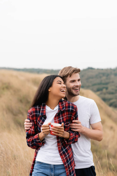Cheerful man hugging african american woman with cup on grassy field during trip  - Photo, Image
