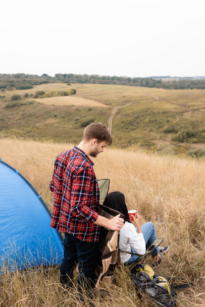 mężczyzna trzyma koc w pobliżu Afryki amerykańskiej dziewczyny z kubkiem i namiotem na trawiastym polu  - Zdjęcie, obraz