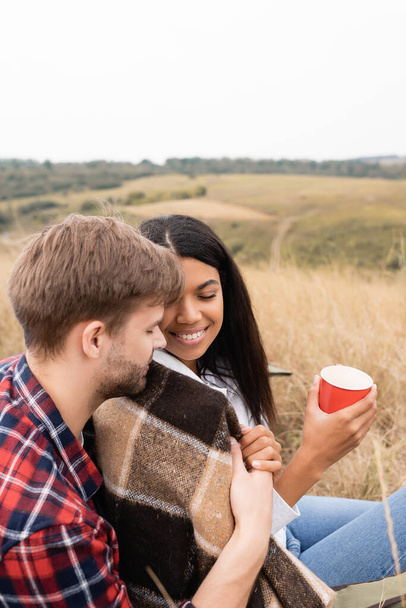 Man hugging smiling african american woman in blanket with cup on grassy meadow  - Foto, afbeelding