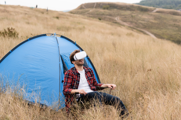 Glimlachende man met behulp van vr headset terwijl zitten in de buurt van tent op het gazon  - Foto, afbeelding