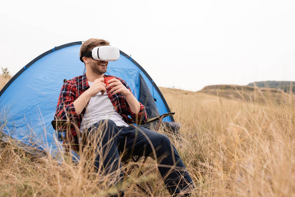 Homme souriant dans vr casque tenant tasse près de la tente sur prairie herbeuse  - Photo, image