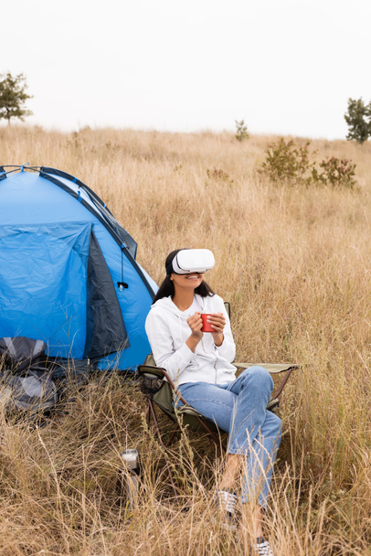 Smiling african american woman in vr headset holding cup near tent on grassy lawn - Photo, Image