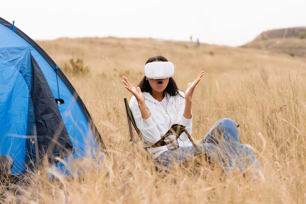 Femme afro-américaine excitée utilisant un casque vr près de la tente sur prairie herbeuse  - Photo, image