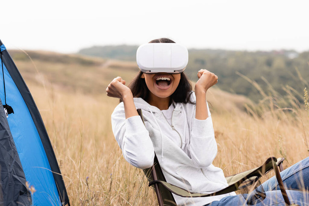 Femme afro-américaine joyeuse utilisant un casque de réalité virtuelle sur une chaise près de la tente sur le terrain  - Photo, image