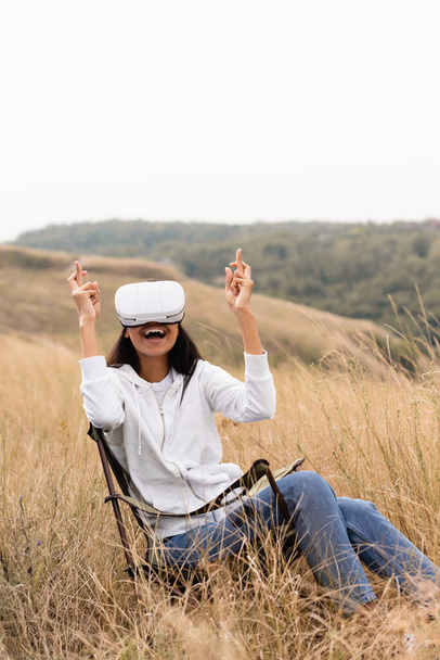 Cheerful african american woman with crossed fingers using  vr headset on chair in field  - Foto, Imagem