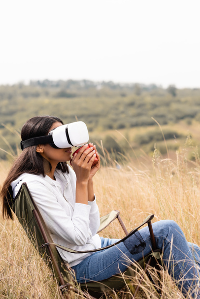 African american woman holding cup while using vr headset on chair and grassy meadow  - Фото, зображення