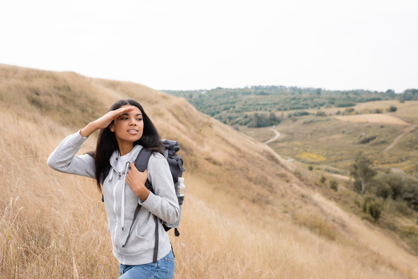 Mosolygó afro-amerikai nő kezében hátizsák és elnézett utazás közben a dombon  - Fotó, kép