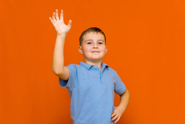 Portarait de caucásico feliz niño sonriente con la mano levantada en la pared del estudio naranja - Foto, imagen