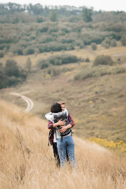 Young man hugging girlfriend with backpack during trip on hill  - Фото, изображение