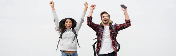 Cheerful multiethnic couple with backpacks and smartphone isolated on grey, banner  - Fotoğraf, Görsel