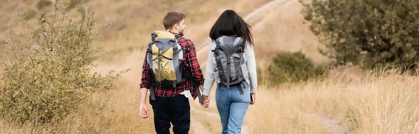 Vista posterior de la joven pareja de viajeros con mochilas caminando en el camino al aire libre, pancarta - Foto, Imagen