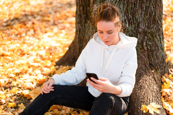 Una donna con un maglione bianco con cappuccio si siede a terra nel parco e tiene in mano un cellulare. carino ragazza uso smartphone per la comunicazione femminile sms un messaggio Communicaion concept  - Foto, immagini
