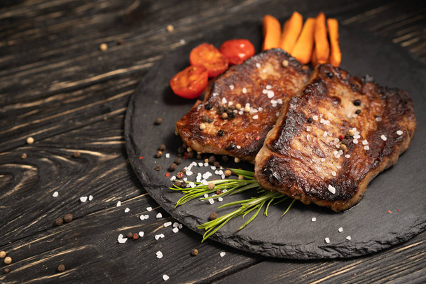 A juicy piece of fried meat with grilled cherry tomatoes and carrot lies on a stone plate against a black wooden table. Degree of roasting well done. Food concept with copy space - Photo, Image