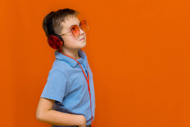 attracive young kid wearing red glasses is listening music with red headphones on orange studio background - Foto, imagen