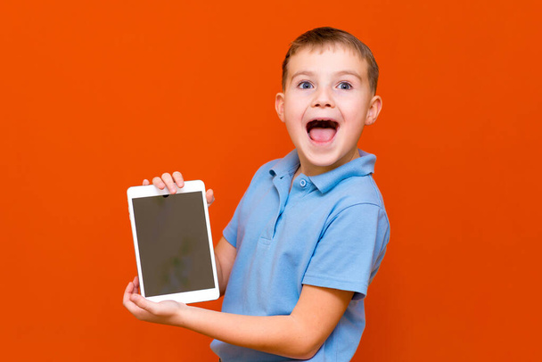 Portrét překvapeného školáka ukazuje tablet a modré tričko na oranžovém pozadí studia.Vesele si něco myslí a ukazuje něco na prázdné obrazovce tablety.oranžové pozadí.Kopírovat prostor, banner. - Fotografie, Obrázek