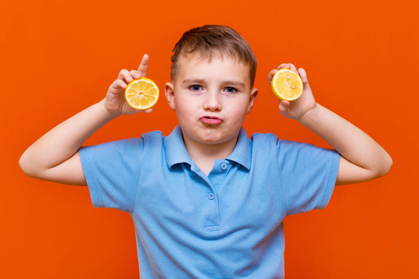 Close up Kaukasisch jong kind toont rauwe schijfjes citroen op een oranje achtergrond. Gezond, leefstijl en een gelukkig kinderconcept. - Foto, afbeelding