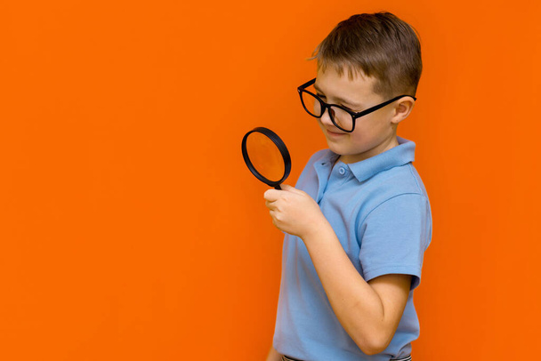 Side view Beautiful european young boy with glasses holding and looking through magnifying glass.Orange studio wall. - Fotoğraf, Görsel