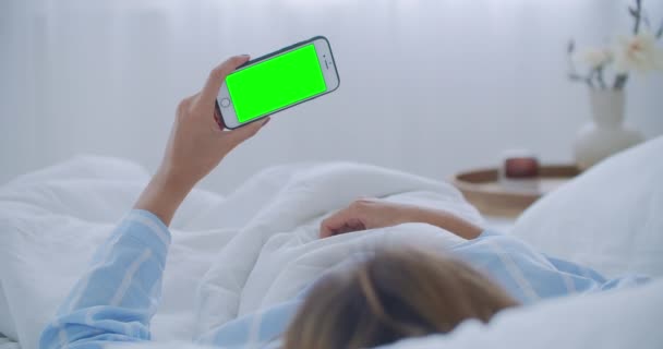 Mujer joven con teléfono celular en la cama, vista al hombro. Adicción a la tecnología. Teléfono de pantalla verde. Mujer joven tocando el vientre y el uso de smartphone en el dormitorio - Metraje, vídeo