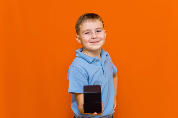 Mały chłopiec w koszulce trzymający czarny prezent dla dziewczynki lub matki, Walentynki, Dzień matki, pomarańczowa ściana studyjna. - Zdjęcie, obraz