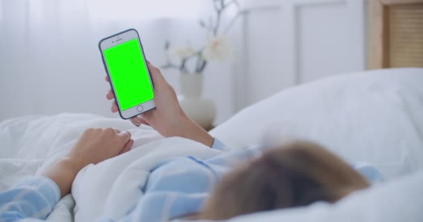 Mujer joven con teléfono celular en la cama, vista al hombro. Adicción a la tecnología. Teléfono de pantalla verde. Mujer joven tocando el vientre y el uso de smartphone en el dormitorio - Imágenes, Vídeo