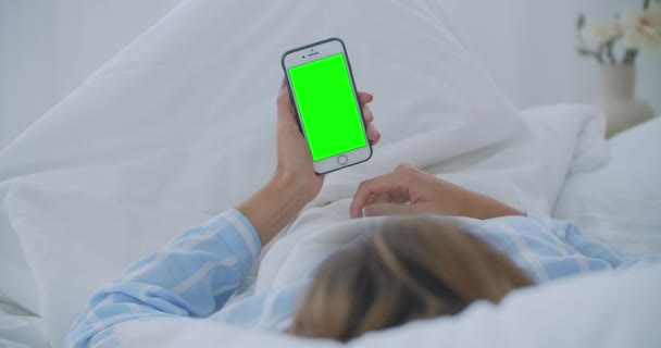 Молодая женщина с мобильным телефоном на кровати, вид на плечо. Технологическая зависимость. Зеленый экран телефона. Молодая женщина трогает живот и использует смартфон в спальне - Кадры, видео